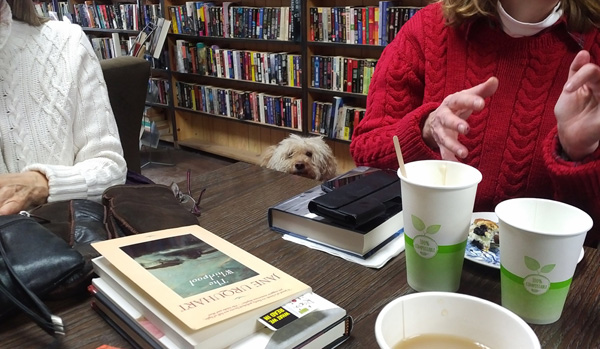 Silent book club meeting with attendees, books, coffee and bookstore dog peeking over table, with bookshelves in background