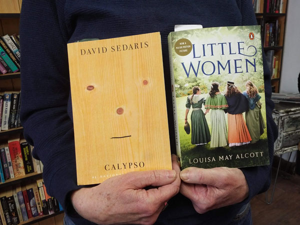 Silent book club participant holds the books Calypso by David Sedaris and Little Women by Louisa May Alcott