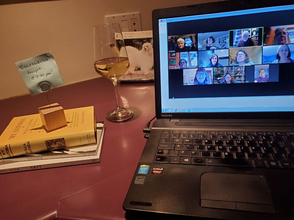 Silent book club meeting shown on computer screen, with stack of book and glass of wine next to computer
