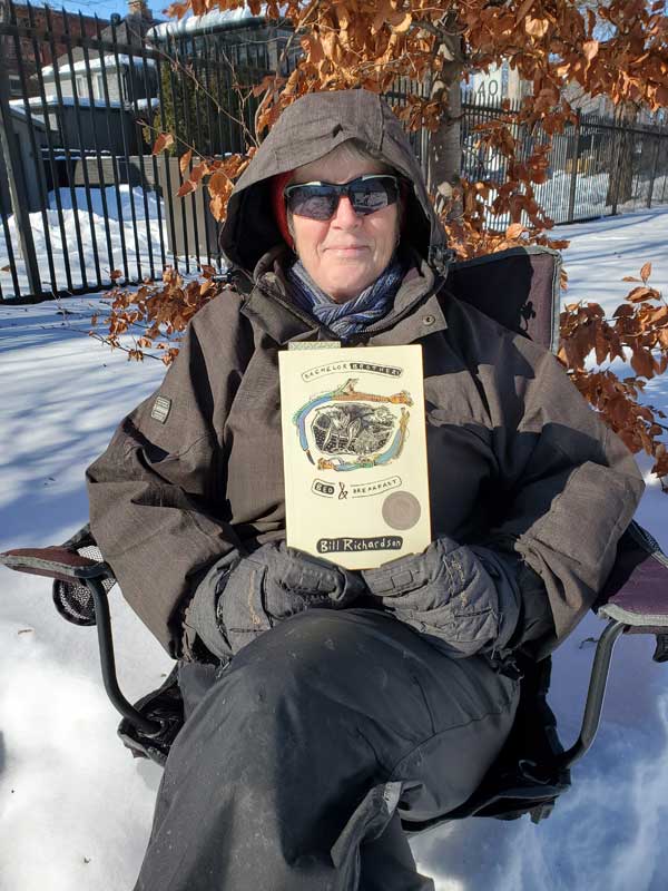 Silent book club member Sue sitting in a chair in snowy Stephenson Park, holding a book by Bill Richardson