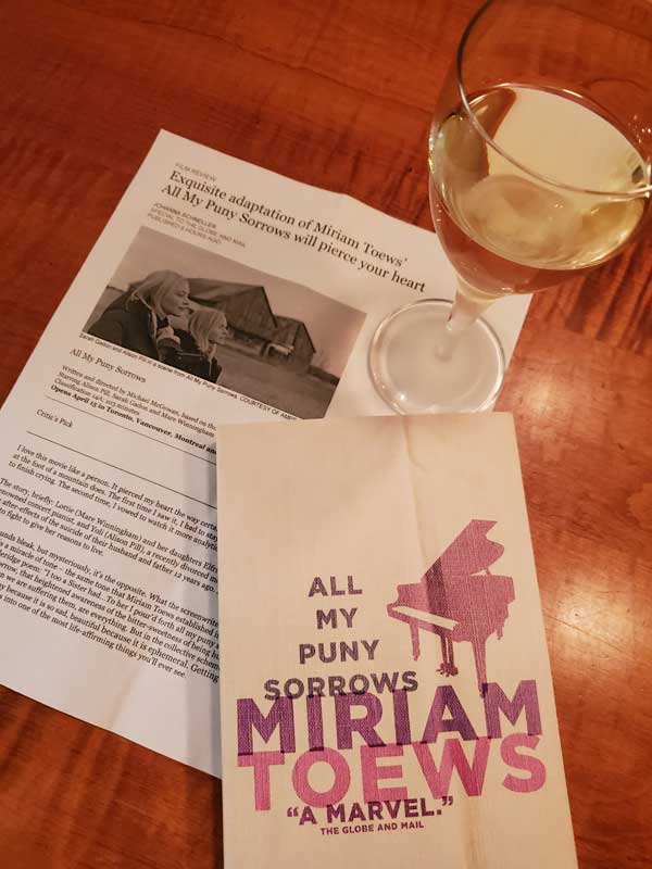The book All My Puny Sorrows by Miriam Toews sits on a table with a print-out of a movie review and a glass of white wine
