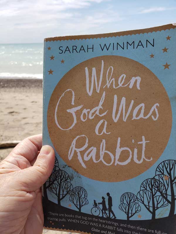 Beth holds up When God Was a Rabbit by Sarah Winman while at the beach