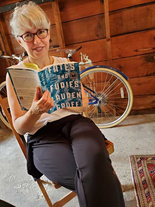 Silent book club member Catherine sits in a wooden chair reading Fates and Furies by Lauren Groff. Her bicycle is leaned up against the wooden wall of the garage in the background.