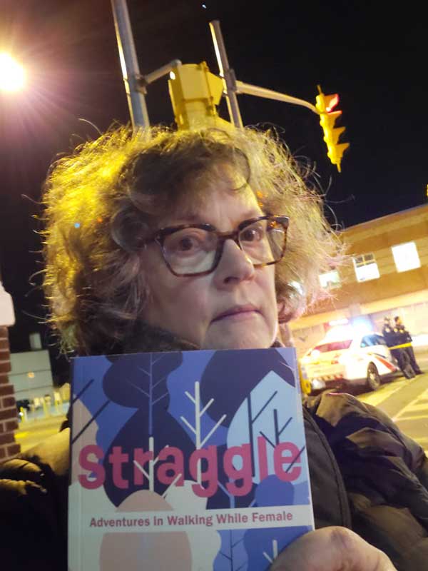 I'm standing on the Danforth at night, under streetlights, with a police cordon in the background, holding Tanis MacDonald's book Straggle - Adventures in Walking While Female