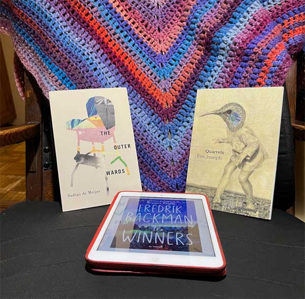 Silent book club member Jo's books, including titles by Sadiqa de Meijer and Eve Joseph - poetry collections in print - and Fredrik Backman, a novel in ebook format, are arranged next to a blue, purple and orange crocheted shawl