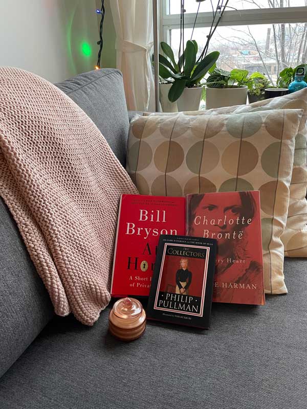 Silent book club member Lyla's books, including titles by Bill Bryson, Claire Harman and Philip Pullman, are arranged on a lovely gray sofa with attractive cushions and afghan, next to a window with house plants on the ledge