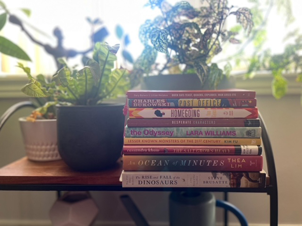Jenn and Sven's books are piled in a sunny, plant-filled window - titles include Post Office by Charles Bukowski, An Ocean of Minutes by Thea Lim and several more.
