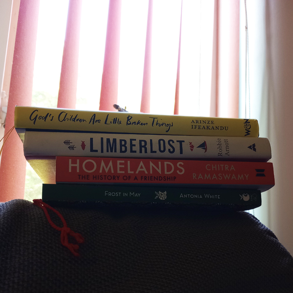 Kathryn's books are piled on the back of a chair, in front of a window with the blinds open - titles include God's Children Are Little Broken Things by Arinze Ifeakandu, Limberlost by Robbie Arnott and more.