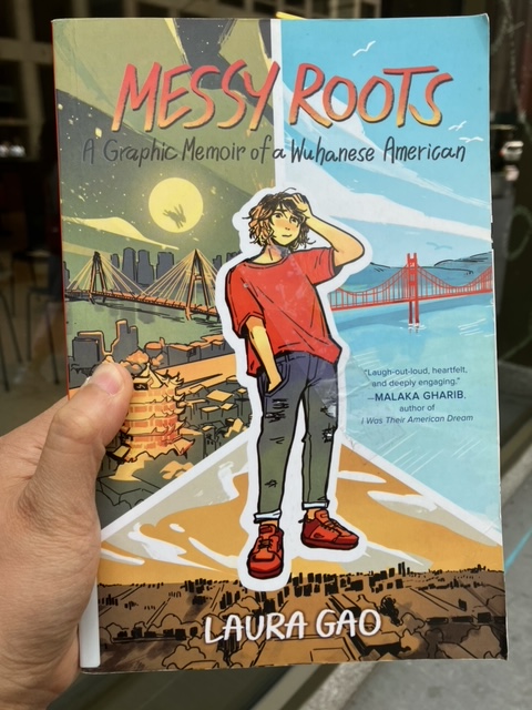 Catherine holds up the graphic memoir Messy Roots by Laura Gao, with a colourful cover showing a drawing of a rueful young woman in jeans and a red T-shirt