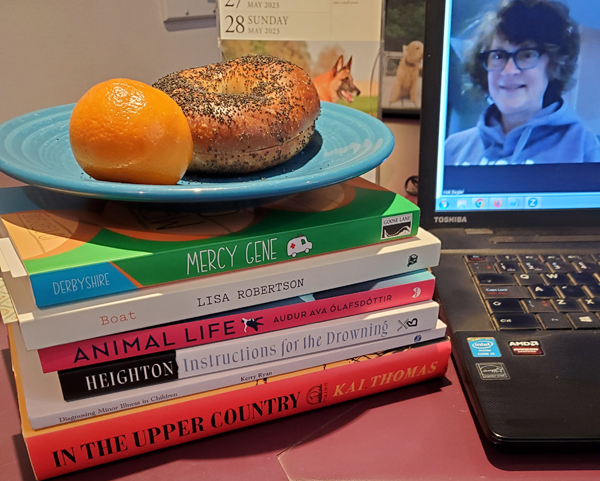 Vicki is on her computer screen, getting ready for a silent book club zoom meeting, with her books piled next to the computer, along with an orange and a bagel - titles include Boat by Lisa Robertson, Mercy Gene by JD Derbyshire and several more.