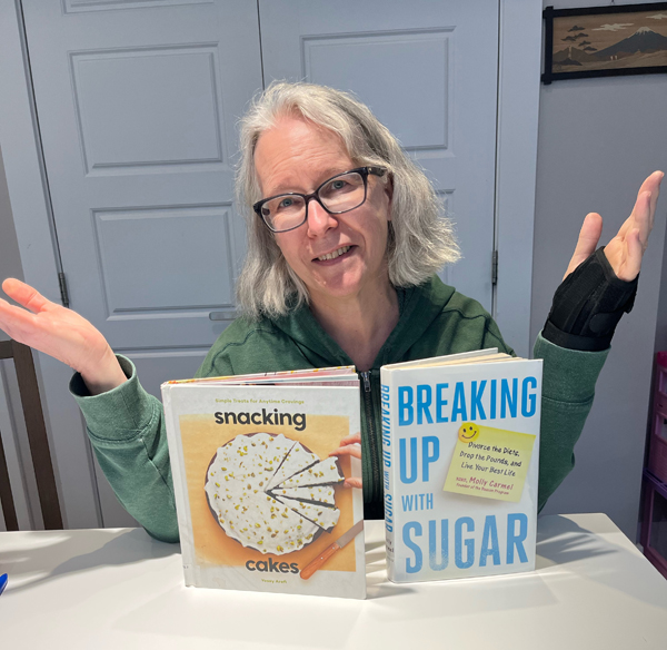 Silent book club member Dawn, with shoulder length silver hair and handsome reading glasses, wearing a green hoodie, gestures ruefully with two books on the white table before her: Snacking Cakes and Breaking Up With Sugar