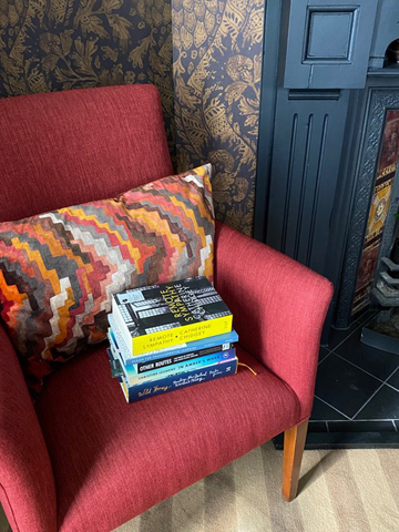 New Zealand silent book club member Marilyn's books are stacked on the seat of a lovely red/pink chair with a red and gold multi-hued cushion. Books include Wild Honey - Reading New Zealand Women's Poetry by Paula Green
