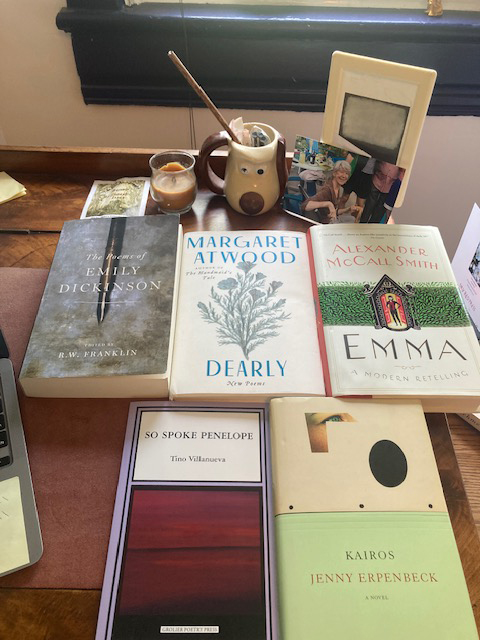 Books arranged on a desk, with a Snoopy mug, including Dearly by Margaret Atwood, Emma by Alexander McCall Smith and more