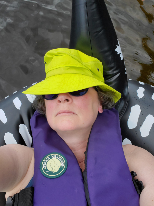 Silent book club member Vicki leans back in an inflatable loon, wearing a life vest, a green neon Tilley hat and sunglasses, contemplating how to read in such a comfy setting