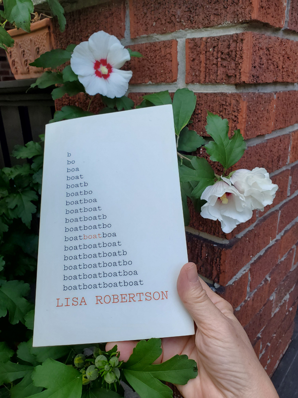 Poetry work Boat by Lisa Robertson, held up near a red brick pillar and some white rose of sharon blossoms