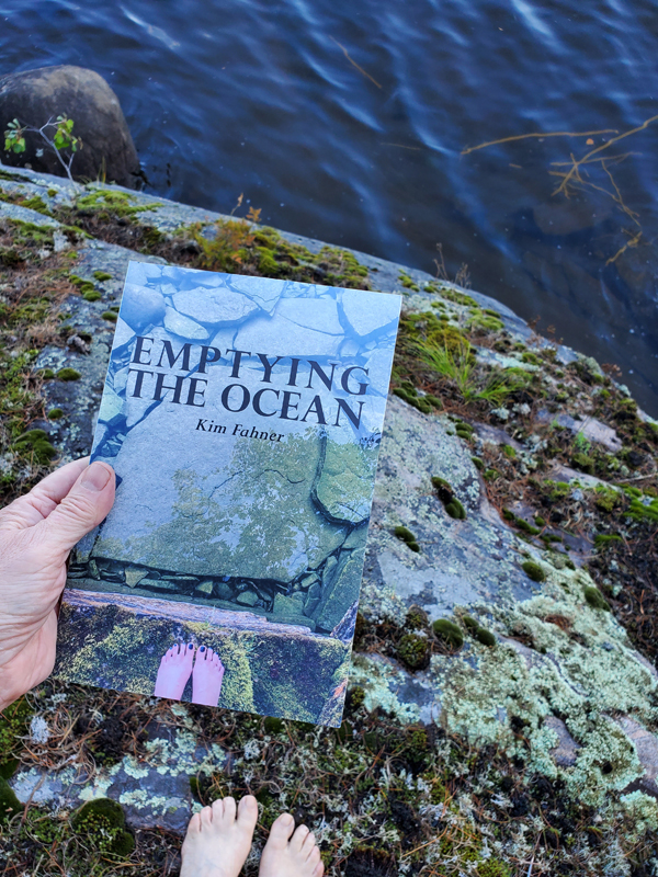 Poetry work Emptying the Ocean by Kim Fahner, held above a mossy, rocky bank looking down toward water, with my bare feet visible, echoing bare feet on the book cover