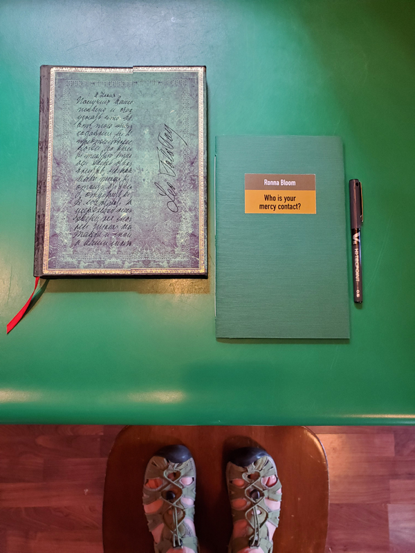 Poetry work Who is Your Mercy Contact? by Ronna Bloom, with a bright green cover, on a bright green counter top with a notebook with an ornate green cover, with my feet in green sandals visible