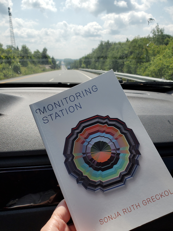 Poetry work Monitoring Station by Sonja Ruth Greckol, held up against a car dashboard as trees and a cell tower are visible through the car window