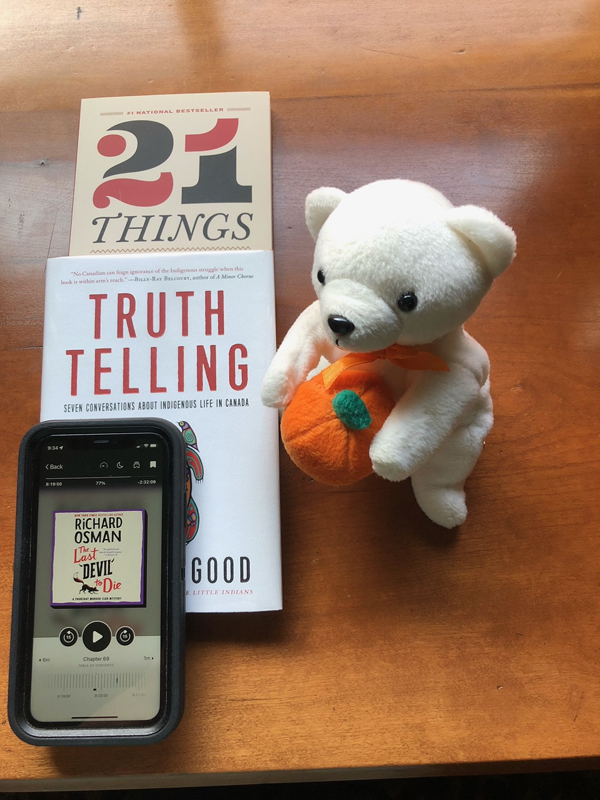 Silent book club member Anne-Louise's recent and current reading, including 21 Things You May Not Know About the Indian Act by Bob Joseph, Truth Telling by Michelle Good and The Last Devil to Die by Richard Osman e-book, accompanied by a white polar bear toy wearing an orange ribbon and holding a plush pumpkin (Photo by Anne-Louise Gould)