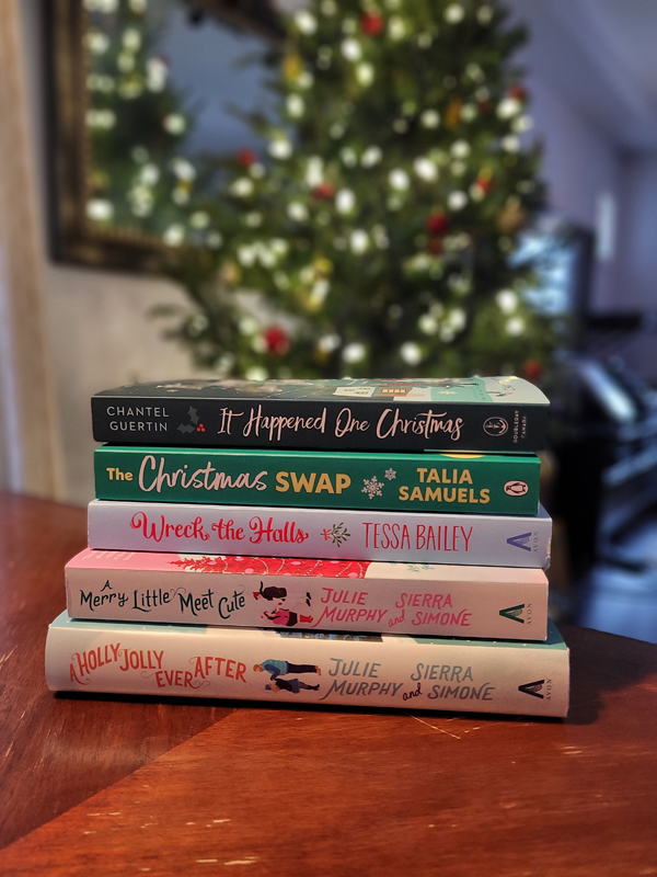 Silent book club member Amber's stack of holiday-themed books, including A Holly Jolly Ever After by Julie Murphy and Sierra Simone. A Christmas tree and lights are visible, blurred, in the background.
