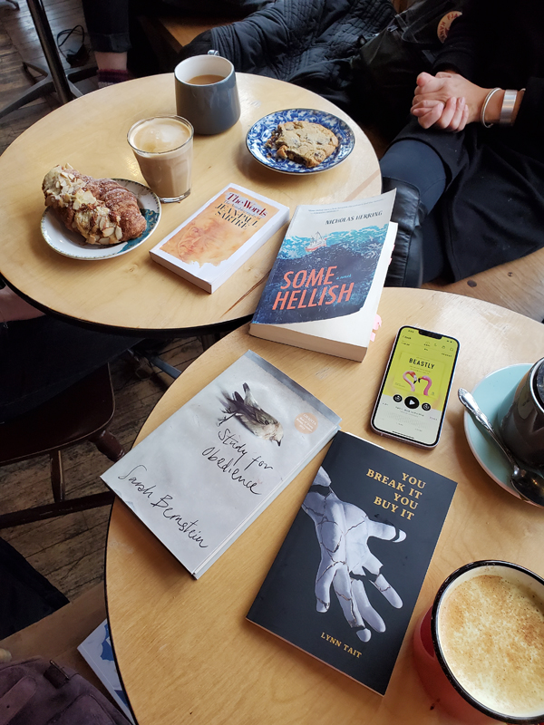 Silent book club meeting at Poured Coffee on Danforth Avenue in Toronto - tables on which books, pastries and coffee/beverages are visible, including Some Hellish by Nicholas Herring, You Break It You Buy It by Lynn Tait and more
