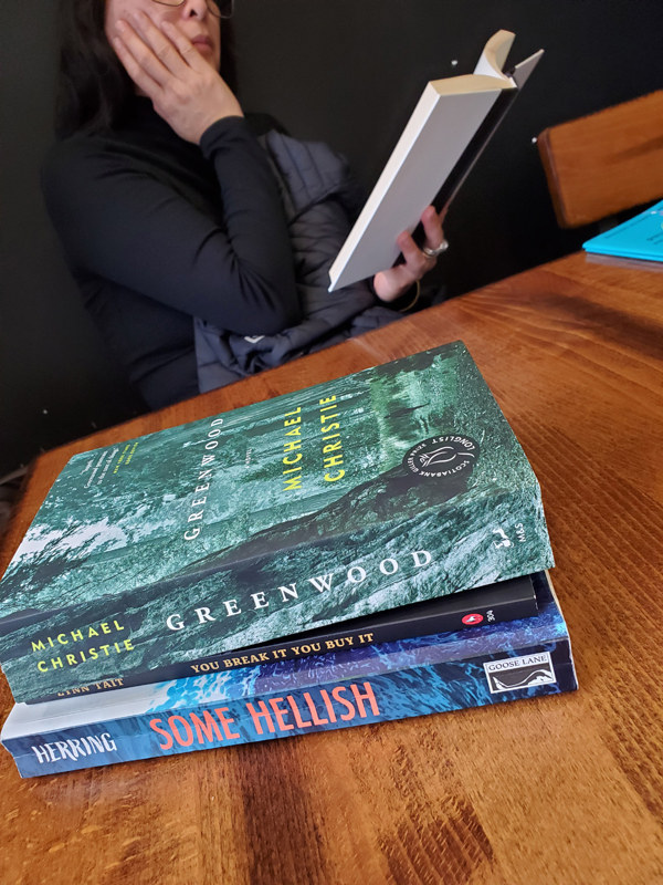 A reader is engrossed in her reading at a silent book club meeting at East Toronto Coffee Co. A stack of books in front of her on the table includes Greenwood by Michael Christie.
