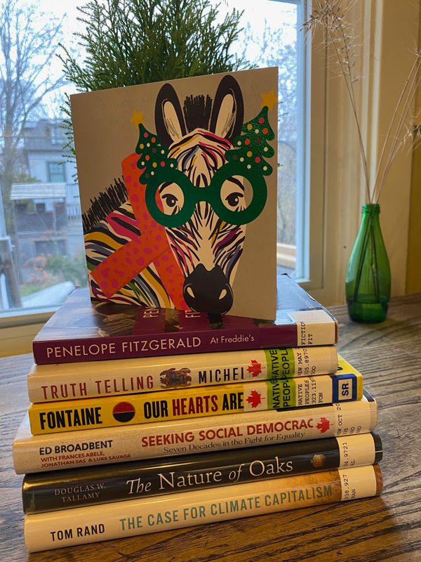 Silent book club member Anne-Louise's stack of books sit in front of a window with a green glass vase nearby, with a holiday card with a cartoon donkey sitting on top of the books. Books include titles by Penelope Fitzgerald, Ed Broadbent, Tom Rand and more.
