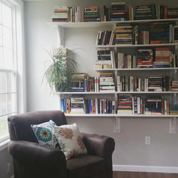 Silent book club member Lisa's beautiful bookshelves, white shelves against a white wall, with a spider plant at one end. Next to the bookshelves are a bright window and a comfy dark leather armchair with cushions with bright floral patterns.