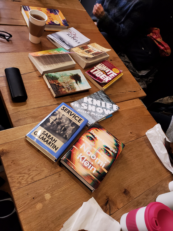 Wooden tables at East Toronto Coffee Co, covered with books by Naomi Klein, Alice Major, Sarah Gilmartin, Tess Gunty, Lisa Elliott, Jodi Picoult, Aravind Adiga, Thomas Mann and more. Readers hands and arms are visible, as are their eyeglasses, beverages and pastries sitting on the tables.
