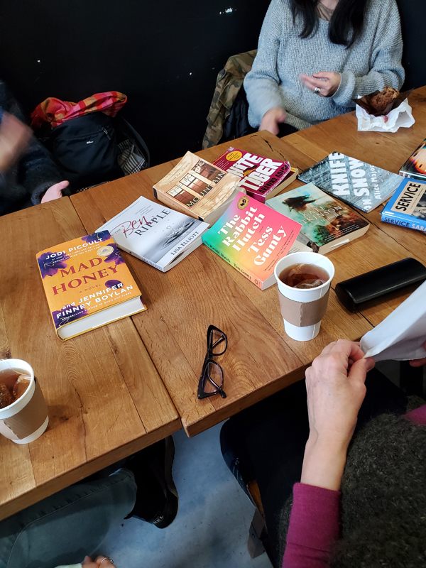 Wooden tables at East Toronto Coffee Co, covered with books by Naomi Klein, Alice Major, Sarah Gilmartin, Tess Gunty, Lisa Elliott, Jodi Picoult, Aravind Adiga, Thomas Mann and more. Readers hands and arms are visible, as are their eyeglasses, beverages and pastries sitting on the tables.