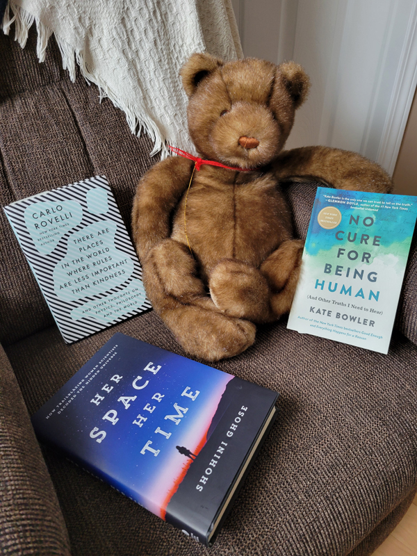 Silent book club member Lori's books - including works by Carlo Rovelli, Kate Bowler and Shohini Ghose - are gently accompanied by a beautiful teddy bear, all curled up in a cozy chair [Photo by Lori]