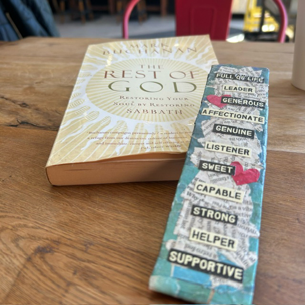 Silent book club member Ruth's reading - The Rest of God by Mark Buchanan - sits on a wooden table with a colourful bookmark with complimentary words on it [Photo by Ruth]
