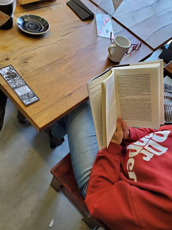 Silent book club member Catherine, wearing a red Dr Pepper sweatshirt, reads at a table at East Toronto Coffee Co - Coffee cups and plates and a bookmark are visible on the table.
