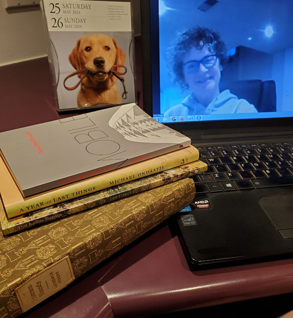 Silent book club member Vicki (that's me, with my signature messy hair and glasses), on screen getting ready for our zoom meeting, with books stacked next to my computer, including work by Michael Ondaatje, Tanis MacDonald, Margaret Christakos and Laurence Sterne