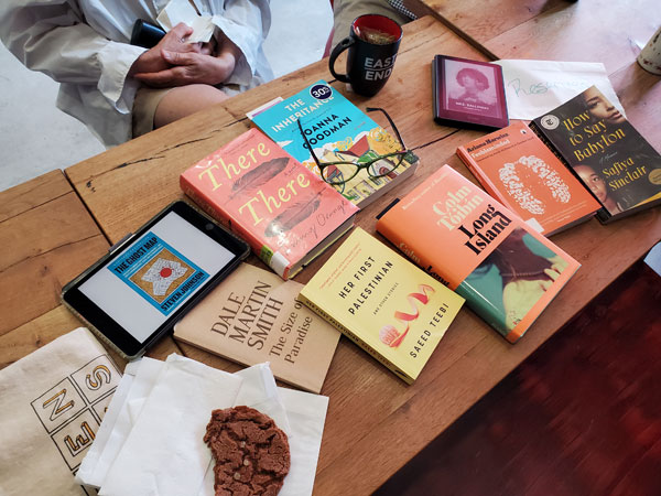 Silent book club meeting at the East Toronto Coffee Co, with the group's book selections spread out on the table along with beverages and pastries. The books in hard cover and digital format include works by Steven Johnson, Dale Martin Smith, Tommy Orange, Saeed Teebi, Joanna Goodman, Colm Toibin, Virginia Woolf, Safiya Sinclair and Ariana Harwicz