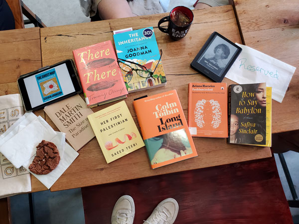 Silent book club meeting at the East Toronto Coffee Co, with the group's book selections spread out on the table along with beverages and pastries. The books in hard cover and digital format include works by Steven Johnson, Dale Martin Smith, Tommy Orange, Saeed Teebi, Joanna Goodman, Colm Toibin, Virginia Woolf, Safiya Sinclair and Ariana Harwicz. My white sneaker-shod feet are visible on the bench next to the table.