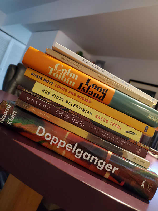 Silent book club member Vicki's colourful stack of recent reading, including books by Naomi Klein, Phil Hall, Pamela Mulloy, Cherie Dimaline, Saeed Teebi, Susie Boyt, Colm Toibin, Dale Martin Smith and Alice Oswald