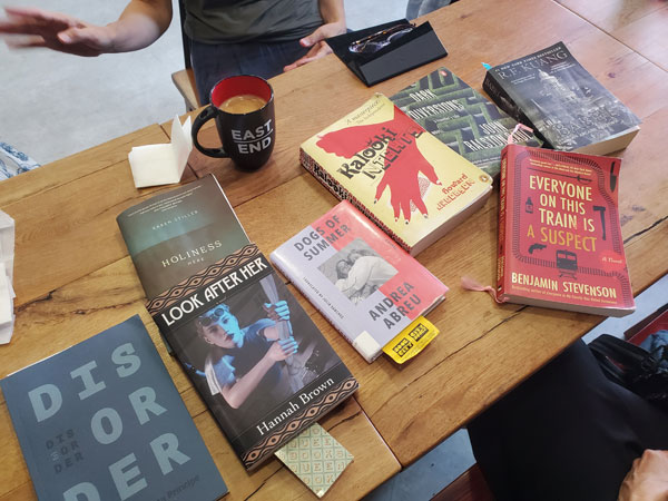 Silent book club meeting at the East Toronto Coffee Co, with the group's book selections spread out on the table along with beverages and pastries. The books include works by Concetta Principe, Hannah Brown, Karen Stiller, Andrea Abreu, Howard Jacobson, Benjamin Stevenson and John Ralston Saul.