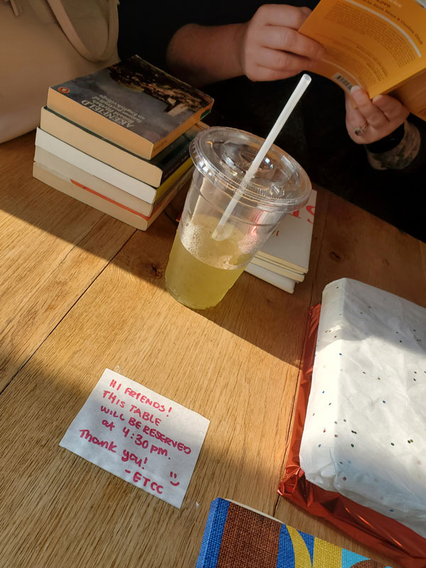 The hands of a reader, holding a book, next to a table laden with more piles of books. The table, at East Toronto Coffee Co, has a 