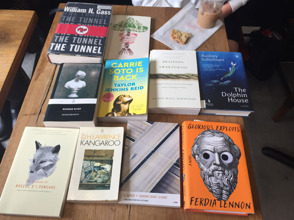 Silent book club meeting at the East Toronto Coffee Co, with the group's book selections spread out on the table along with beverages and pastries. The books include works by D.H. Lawrence, Ferdia Lennon, William Gass, Audrey Schulman and George Eliot.