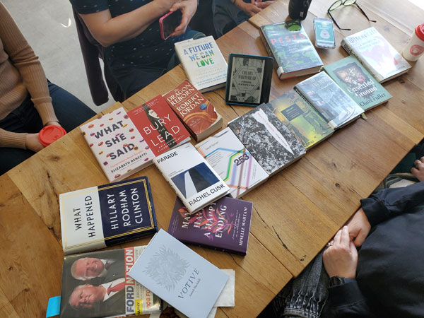 Silent book club meeting at the East Toronto Coffee Co, with the group's book selections spread out on the table along with beverages and pastries. The books include works by Rob Ford and Doug Ford, Hillary Rodham Clinton, Annick MacAskill, Rachel Cusk, Elizabeth Renzetti, Thomas Pynchon, Claire Fuller + more