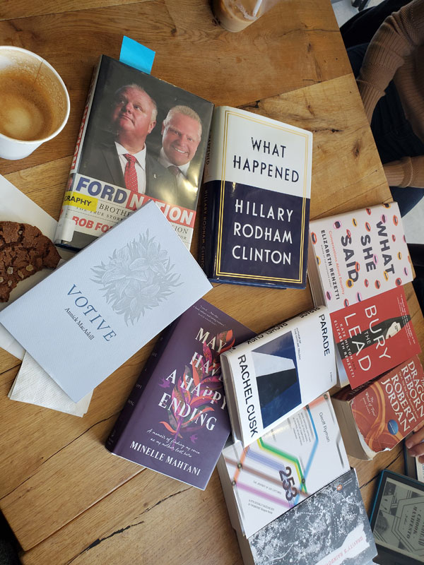 Silent book club meeting at the East Toronto Coffee Co, with some of the group's book selections spread out on the table along with beverages and pastries. The books include works by Rob Ford and Doug Ford, Hillary Rodham Clinton, Annick MacAskill, Rachel Cusk, Elizabeth Renzetti, Geoff Ryman, Robert Jordan + more