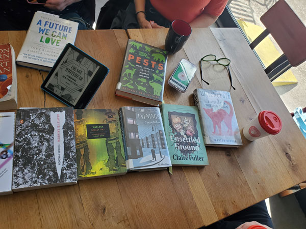 Silent book club meeting at the East Toronto Coffee Co, with some of the group's book selections spread out on the table along with beverages and pastries. The books include works by Thomas Pynchon, Oakley Hall, Claire Fuller, Gerard Reve + more