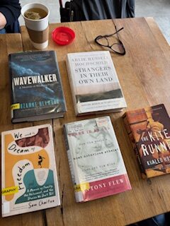 Silent book club members gathered, reading silently, at a table at East Toronto Coffee Co. Here are some of the books they read, on a ETCC table (including a pair of glasses and tea in a takeout cup): Wavewalker: A Memoir of Breaking Free by Suzanne Heywood, Strangers in Their Own Land - Anger and Mourning on the American Right by Arlie Russell Hochschild, We Used to Dream of Freedom - A Memoir of Family, the Holocaust, and the Stories We Don't Tell by Sam Chaiton, There Is a God by Antony Flew and The Kite Runner by Khaled Hosseini