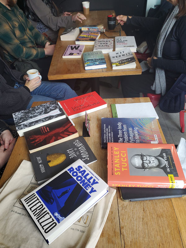 Silent book club meeting at the East Toronto Coffee Co, with the group's book selections spread out on the table along with beverages and pastries. The books include works by Sally Rooney, Stanley Tucci, Jason Heroux, Thomas Pynchon + more