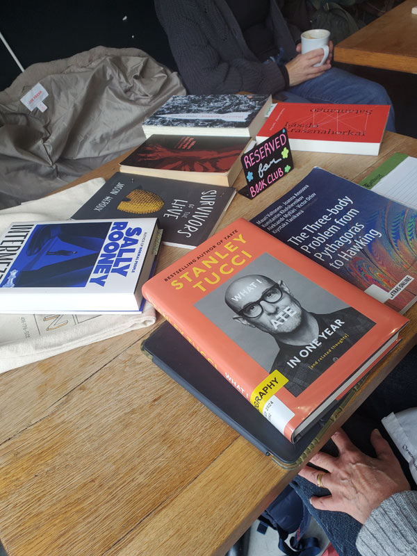 Silent book club meeting at the East Toronto Coffee Co, with some of the group's book selections spread out on the table along with beverages and pastries. The books include works by Sally Rooney, Stanley Tucci, + more