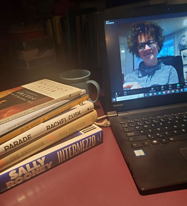 Silent book club member Vicki (that's me, with my signature messy hair and glasses), on screen getting ready for our zoom meeting, with books stacked next to my computer, including works by Sally Rooney, Olga Tokarczuk, Rachel Cusk, Billy Collins and more