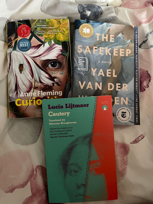 Silent book club member Lyla's book pile: Curiosities by Anne Fleming, The Safekeep by Yael van der Wouden and Cautery by Lucía Lijtmaer, translated by Maureen Shaughnessy