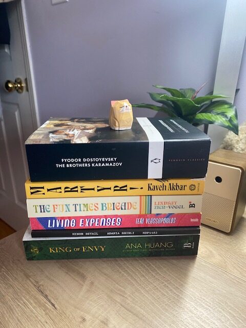 Silent book club member Ghada's book pile: Minor Detail by Adania Shibli, Martyr! by Kaveh Akbar, Living Expenses by Teri Vlassopolou, The Fun Times Brigade by Lindsay Zier-Vogel, King of Envy by Ana Huang, The Brothers Karamazov by Fyodor Dostoevsky