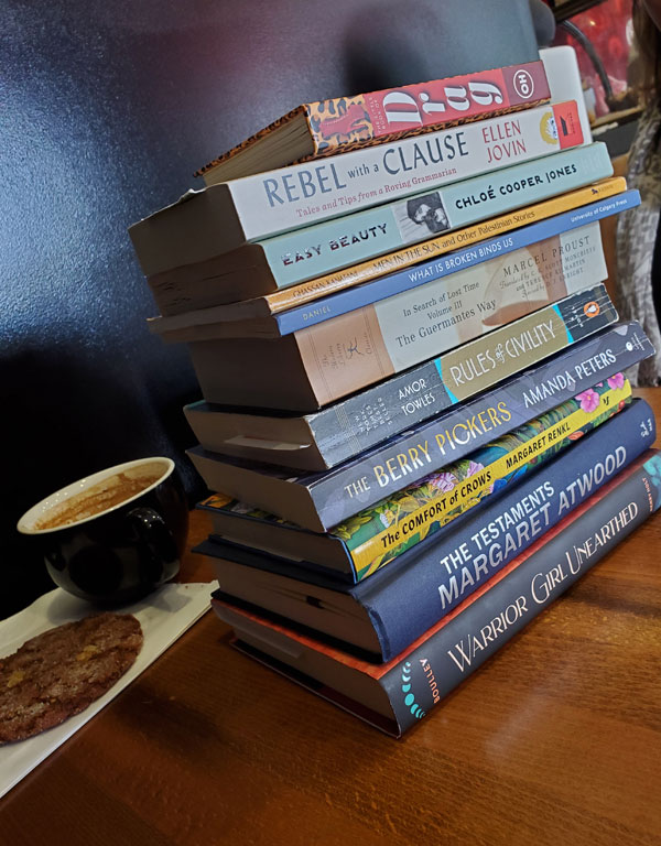 Group book pile at East Toronto Coffee Co, accompanied by coffee - titles include Easy Beauty by Chloe Cooper Jones, The Testaments by Margaret Atwood, The Comfort of Crows by Margaret Renkl + more